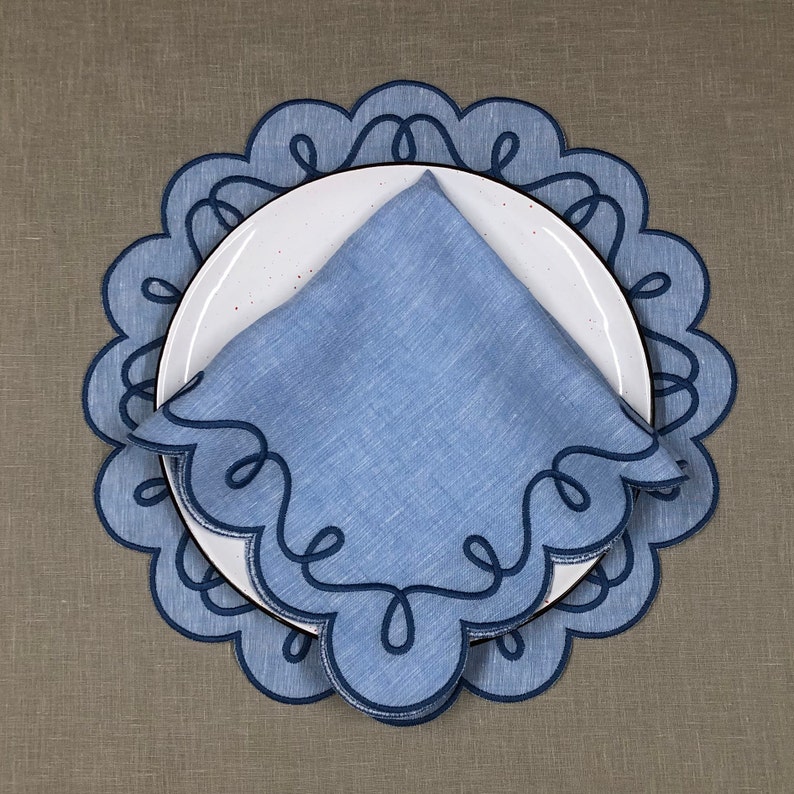 May include: Blue linen napkin with a scalloped edge and a decorative design. The napkin is placed on a white plate with a blue and white scalloped edge.