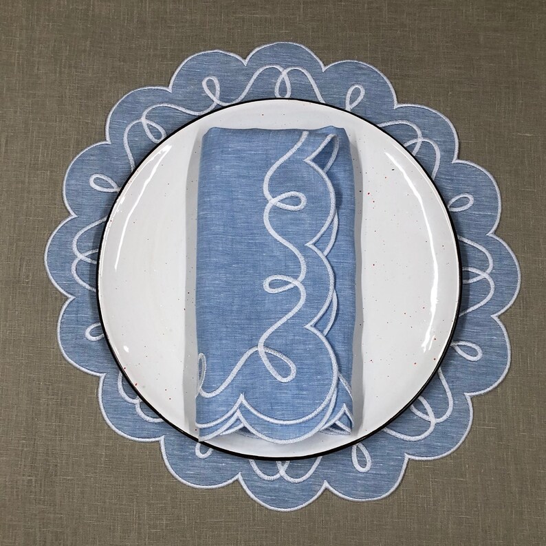 May include: A blue linen napkin with white embroidered scalloped edges, folded and placed on a white plate with a black rim. The plate is on top of a blue and white scalloped placemat.