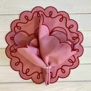 May include: A pink linen napkin with scalloped edges and a red embroidered design. The napkin is folded and placed on a pink and red scalloped placemat.