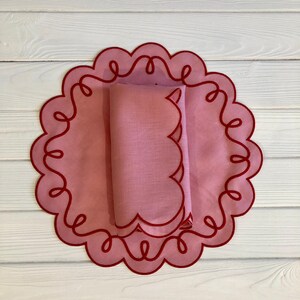 May include: A pink scalloped placemat with a red embroidered border. The placemat is folded in half and placed on top of a second, similar placemat.