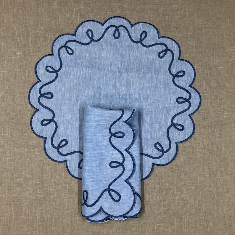May include: Blue linen placemat with a scalloped edge and a matching blue linen napkin. Both have a dark blue embroidered design.