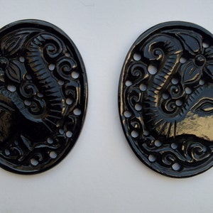 Könnte beinhalten: Zwei schwarze ovale Anhänger mit einem aufwendig geschnitzten Design eines Elefantenkopfes mit floralen Akzenten.