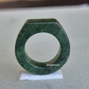 May include: A dark green, faceted ring with a smooth, polished finish. The ring has a geometric design and is made of a solid, opaque material. The ring is displayed on a small, white stand. The text "jadigemsart" is visible on the ring.