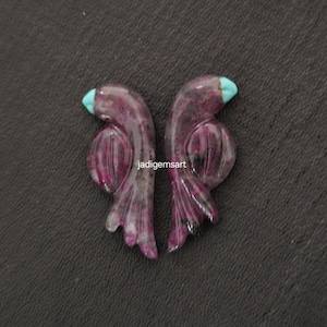 May include: Two carved gemstone bird pendants. The pendants are a deep purple with red and white veining, and turquoise blue accents. The pendants are displayed on a dark gray surface. The text "jadigemsart" is visible.