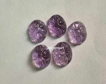 Könnte beinhalten: Fünf ovale Amethyst-Edelsteine mit geschnitzten Blumenmustern. Die Edelsteine haben eine durchscheinende violette Farbe und liegen auf einer hellen Oberfläche. Die geschnitzten Details erzeugen einen dreidimensionalen Effekt.