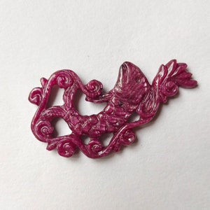 May include: A carved ruby gemstone pendant in the shape of a fish swimming through a vine. The pendant is a deep pink color and has intricate details.