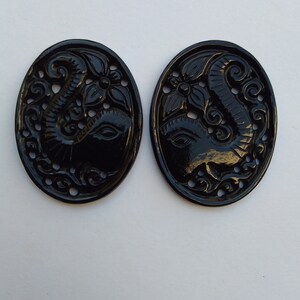 May include: Two black oval-shaped pendants with intricate carvings of an elephant head with floral designs. The pendants are made of a smooth, polished material.