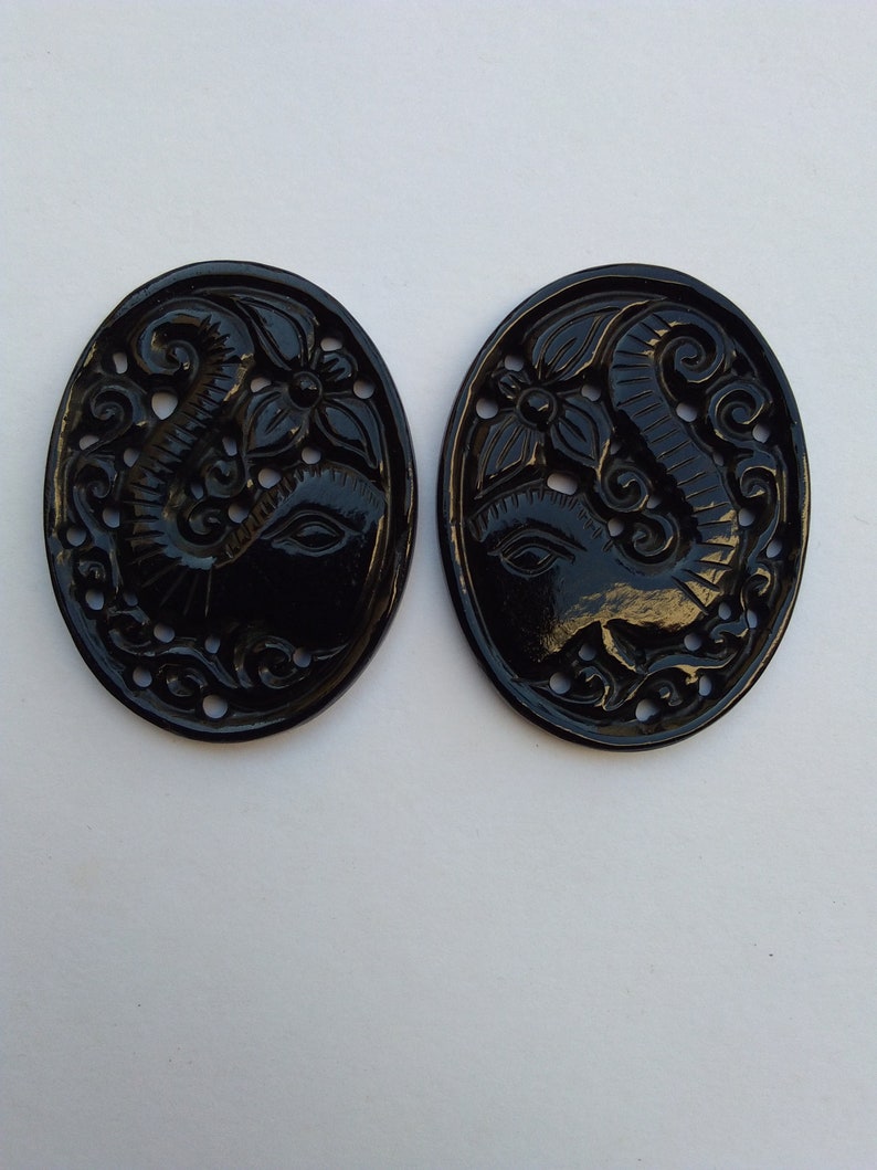 May include: Two black oval-shaped pendants with intricate carvings of an elephant head with floral designs. The pendants are made of a smooth, polished material.