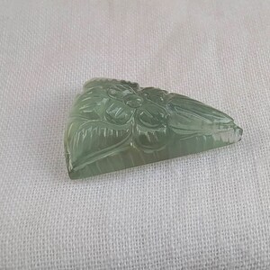 May include: A carved green gemstone pendant with a floral design. The pendant is shaped like a triangle and has a smooth, polished surface.