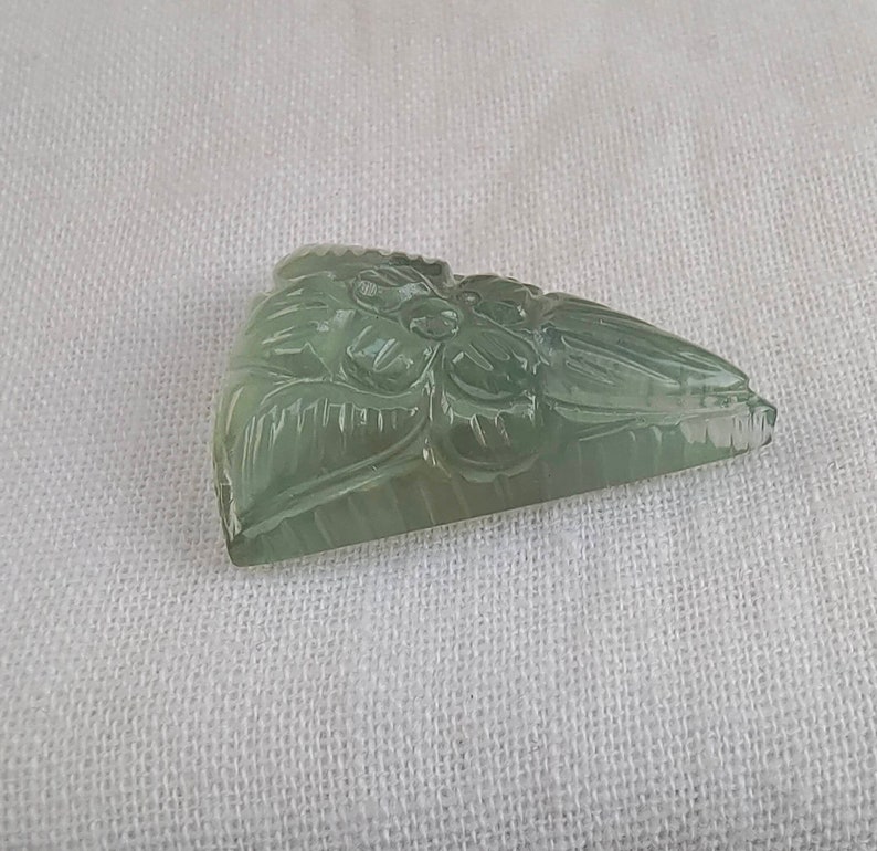 May include: A carved green gemstone pendant with a floral design. The pendant is shaped like a triangle and has a smooth, polished surface.