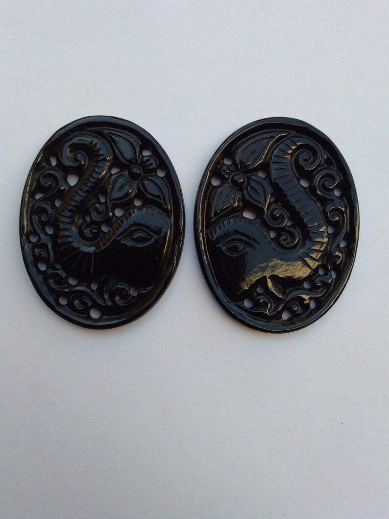 May include: Two black oval-shaped pendants with intricate carvings of an elephant head with floral designs. The pendants are made of a smooth, polished material.