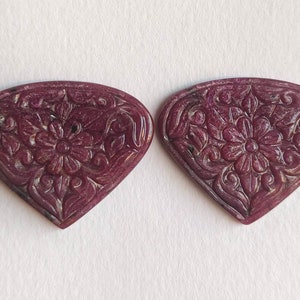 Op de afbeelding: Twee gegraveerde robijnrode hartvormige edelstenen met ingewikkelde bloemmotieven. De edelstenen zijn diep rood van kleur en hebben een gepolijste afwerking.