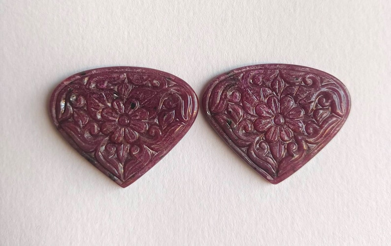 Op de afbeelding: Twee gegraveerde robijnrode hartvormige edelstenen met ingewikkelde bloemmotieven. De edelstenen zijn diep rood van kleur en hebben een gepolijste afwerking.