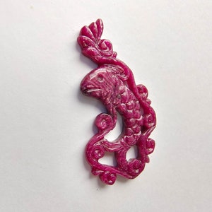 May include: A carved ruby pendant in the shape of a fish with a floral design. The pendant is a deep pink color and has a glossy finish.