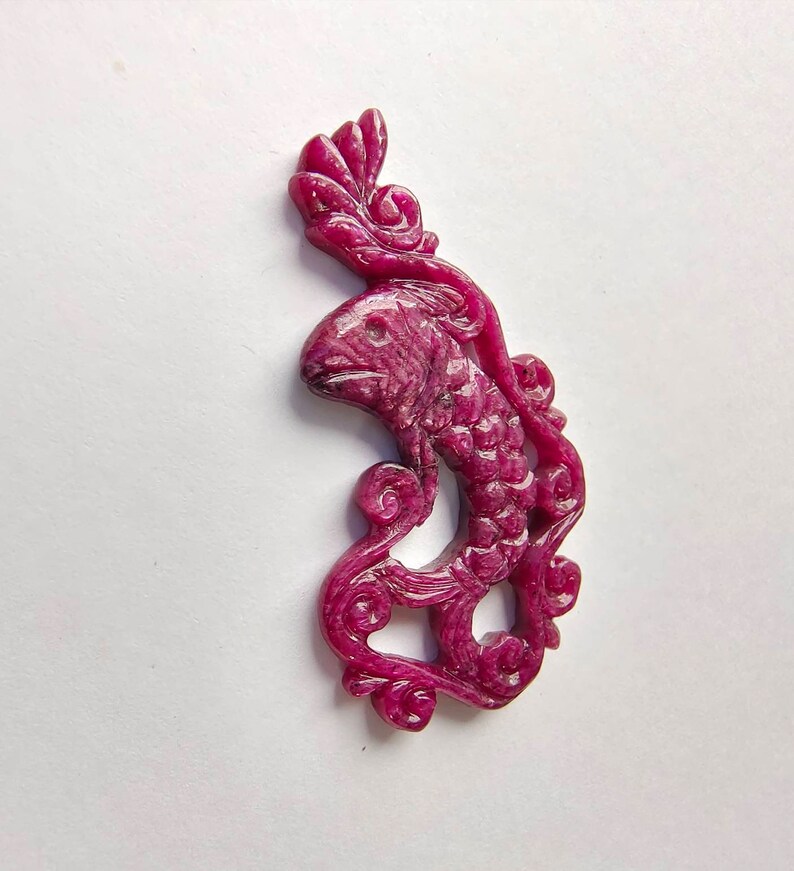 May include: A carved ruby pendant in the shape of a fish with a floral design. The pendant is a deep pink color and has a glossy finish.