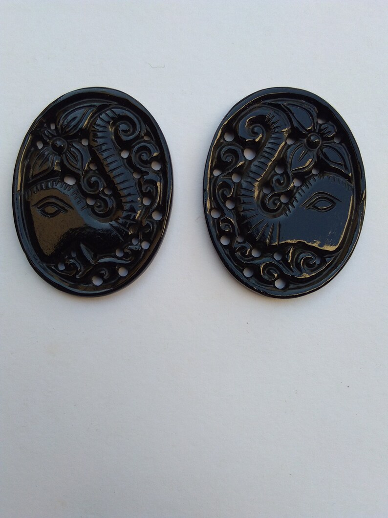 May include: Two black oval-shaped buttons with an intricate carved design of an elephant's head with a floral motif.