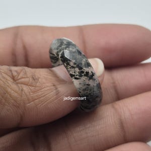 Op de afbeelding: Een facetgeslepen ring met een gespikkeld grijs en zwart patroon. De ring is gemaakt van een stenen materiaal en heeft een gepolijste afwerking. De tekst "jadigemsart" is zichtbaar op de ring.
