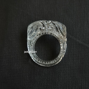 May include: A clear, carved ring with an ornate floral and scroll pattern. The ring is made of a transparent material, possibly crystal or glass, and has a wide band. The ring is displayed against a dark background. The text "jadigemsart" is visible.