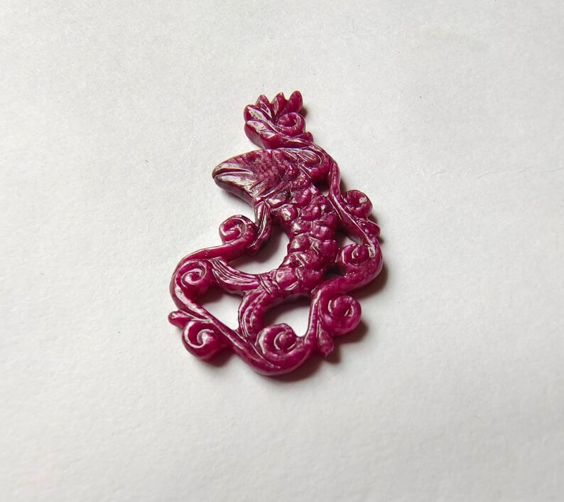 May include: A carved ruby pendant in the shape of a fish with intricate details. The pendant is a deep red color and has a glossy finish.