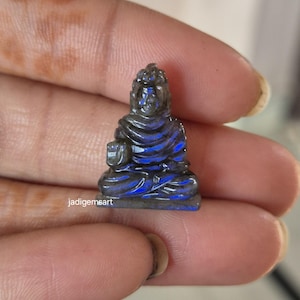 Pode incluir: Pequena estatueta esculpida de um Buda sentado. A escultura é feita de uma pedra escura e iridescente com tons azuis e cinzentos. A figura está em pose meditativa, com as mãos no colo. O texto "jadigemsart" é visível.