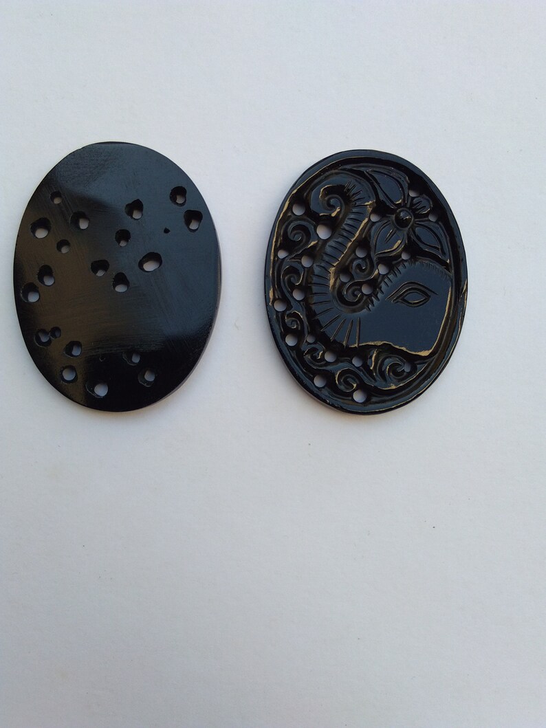 May include: Two black oval-shaped carved buttons. One button has a detailed carving of an elephant with a flower. The other button has a pattern of small circles.