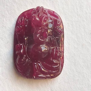 May include: A carved ruby gemstone pendant featuring a detailed depiction of the Hindu deity Ganesha. The pendant is a deep red color and has a smooth, polished finish.