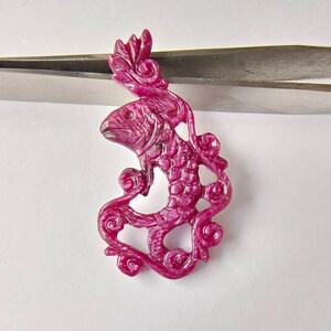 May include: A carved ruby pendant in the shape of a fish swimming through a swirl of vines. The pendant is a deep pink color and has intricate details.