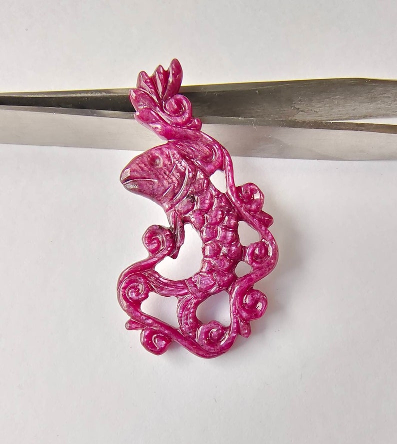 May include: A carved ruby pendant in the shape of a fish swimming through a swirl of vines. The pendant is a deep pink color and has intricate details.