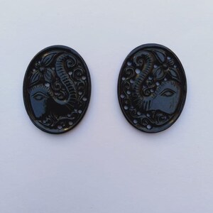 May include: Two black oval-shaped buttons with an intricate carved design of an elephant's head and trunk. The buttons have a glossy finish and are perfect for adding a touch of elegance to any project.
