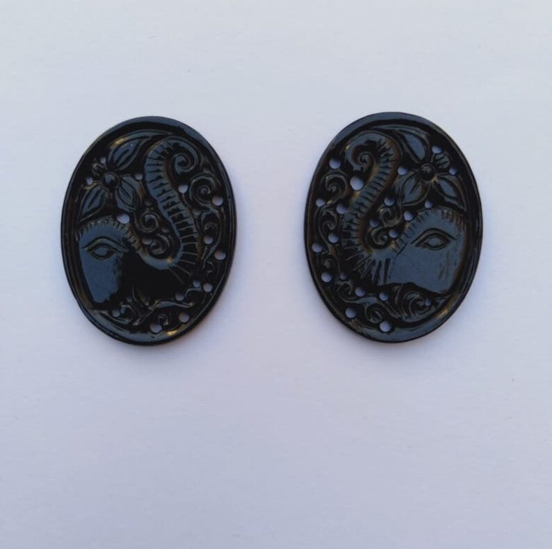 May include: Two black oval-shaped buttons with an intricate carved design of an elephant's head and trunk. The buttons have a glossy finish and are perfect for adding a touch of elegance to any project.