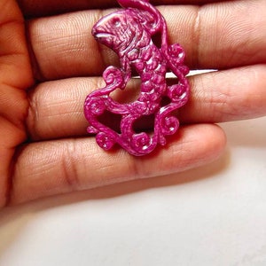 May include: A carved ruby pendant in the shape of a fish with intricate details. The pendant is a deep pink color and has a glossy finish.