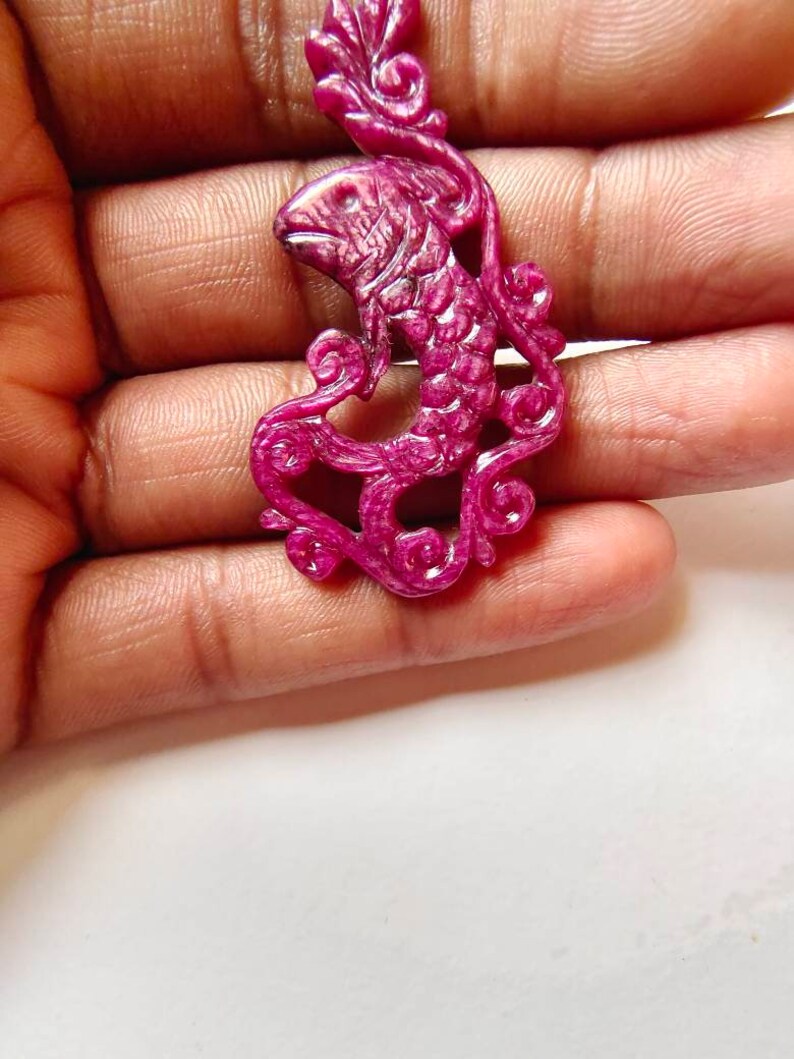 May include: A carved ruby pendant in the shape of a fish with intricate details. The pendant is a deep pink color and has a glossy finish.