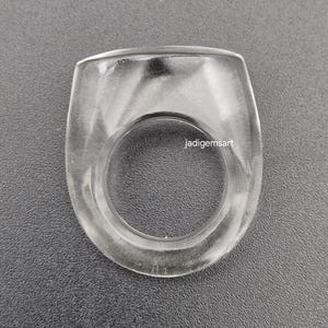 Könnte beinhalten: Ein klarer, transparenter Ring mit rechteckiger Oberseite und kreisförmiger Öffnung. Der Ring besteht aus einem glatten, polierten Material und ist vor einem grauen Hintergrund platziert. Der Text "jadigemsart" ist auf dem Ring sichtbar.