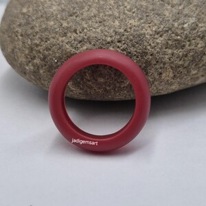 Peut inclure: Une bague de couleur bordeaux avec une finition lisse et mate. La bague est circulaire et repose sur une surface de couleur claire. Le texte "jadigemsart" est imprimé sur la bague.