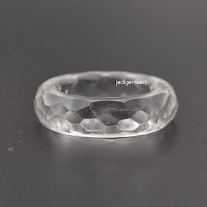 Puede incluir: Un anillo transparente facetado con un diseño geométrico. El anillo está hecho de un material transparente, posiblemente cristal o vidrio, y presenta una serie de cortes angulados en su superficie. El anillo es de estilo banda.