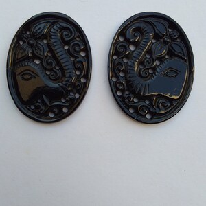 May include: Two black oval-shaped buttons with an intricate carved design of an elephant's head with a floral motif.