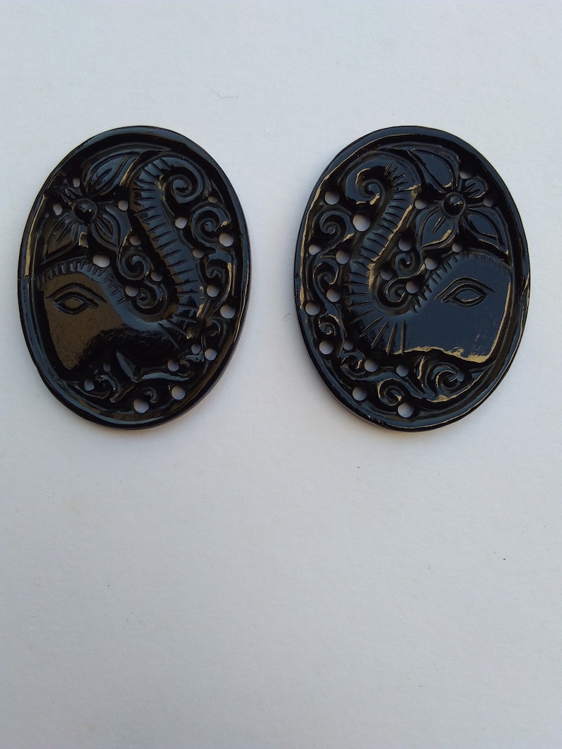 May include: Two black oval-shaped buttons with an intricate carved design of an elephant's head with a floral motif.