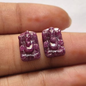 May include: Two carved ruby gemstones, each depicting a deity, are displayed. The gemstones are rectangular with a deep pink hue. The text "jadigemsart" is visible on each stone. The carvings are detailed and show the form of the deity.