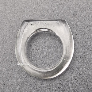 Peut inclure: Une bague transparente et claire avec un anneau arrondi et un dessus plat et rectangulaire. La bague est faite d'un matériau lisse et poli. Le design est simple et élégant. La bague est présentée sur une surface grise.