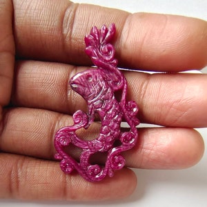 May include: A carved ruby pendant in the shape of a fish with intricate floral details. The pendant is a deep pink color and has a glossy finish.
