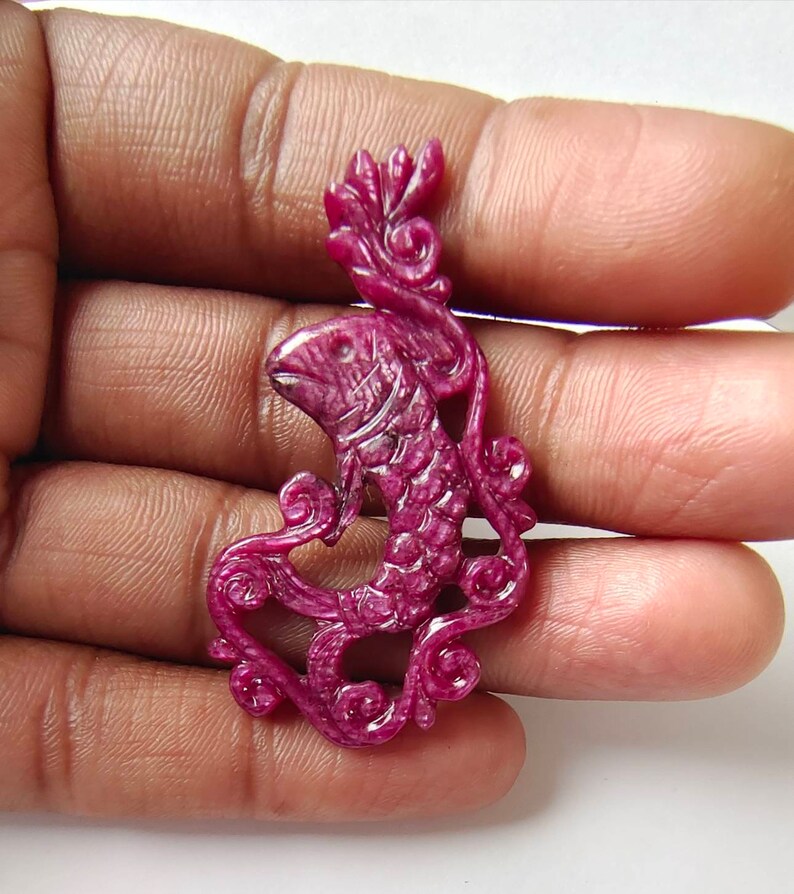May include: A carved ruby pendant in the shape of a fish with intricate floral details. The pendant is a deep pink color and has a glossy finish.