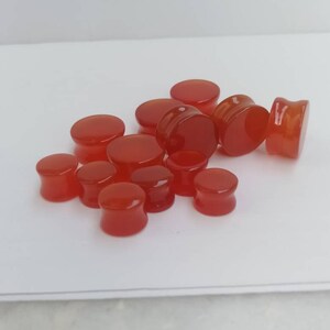 May include: A collection of 12 red carnelian stone ear gauges, also known as plugs or tunnels, for stretched earlobes. The gauges are smooth and polished, with a natural carnelian color.
