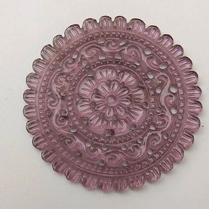 Puede incluir: Un adorno redondo de vidrio morado con intrincados diseños tallados. El adorno tiene un patrón floral en el centro y un borde festoneado.