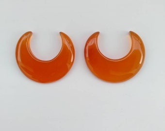May include: Two orange crescent moon shaped cabochons, likely made of agate, on a white background.