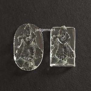 Könnte beinhalten: Zwei klare Kristallanhängern mit einer geschnitzten Darstellung einer hinduistischen Gottheit, die Flöte spielt. Die Anhänger sind oval und rechteckig.