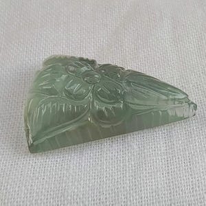 May include: A carved green jade pendant featuring a floral design. The pendant is triangular in shape and has a smooth, polished surface.
