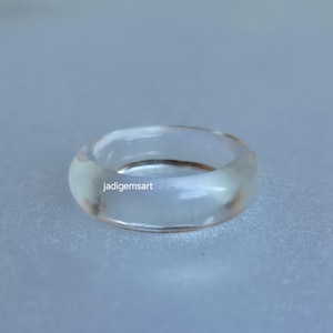 May include: A clear, transparent ring with a rounded shape. The ring appears to be made of a smooth, polished material. The text "jadigemsart" is visible on the ring. The ring is a piece of jewellery.