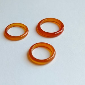 May include: Three orange carnelian rings, each with a smooth, polished finish. The rings are stacked on a white surface.