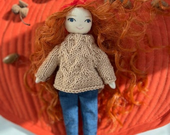 May include: A handmade doll with red yarn hair and blue eyes, wearing a tan knit sweater and blue jeans. A red ribbon adorns the doll's hair. The doll is set against an orange pumpkin-shaped background with acorns.