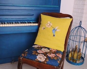 May include: A yellow throw pillow with a floral embroidery design featuring a hummingbird. The pillow is on a chair with a floral patterned fabric. A blue piano is in the background.